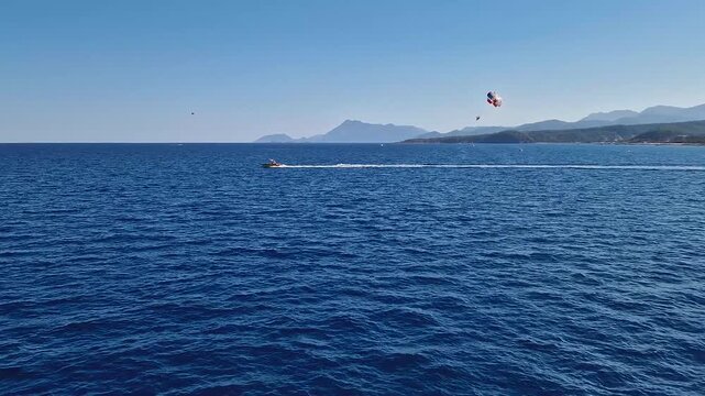 Seascape parasailing adventure over the vast, blue sea on the Turkish Mediterranean coast. Speedboat pulls a parasailer, creating a white wake on the water. Majestic Taurus mountains range at horizon