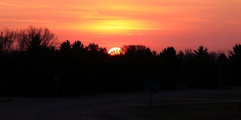 A vibrant sunset over a dark treeline with a glowing sky and a hint of clouds at the horizon line
