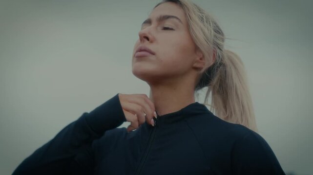 A female in athletic clothing, zipping up near the beach or sea under an overcast sky.