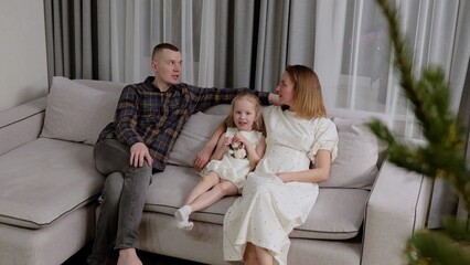 Pregnant mom, dad, and daughter relaxing together on living room sofa, sharing warm family moment and anticipating new arrival