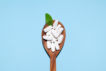 White tablets, vitamins, pills and green leaf in wooden spoon on blue background top view. Medical pharmaceutical background, health concept, organic dietary supplements, self care