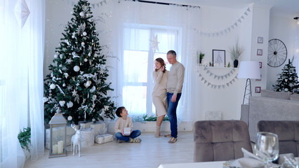 A day with family before Christmas. New Year's Eve with family. Young boy holding a gift while his parents stand next to a christmas tree in a festively decorated living room
