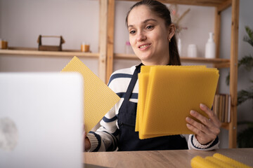 happy woman showing beeswax sheets for handmade candles during video call on laptop in craft workshop.