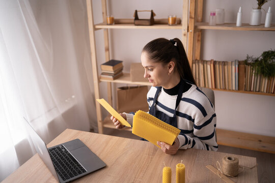 happy woman showing beeswax sheets for handmade candles during video call on laptop in craft workshop