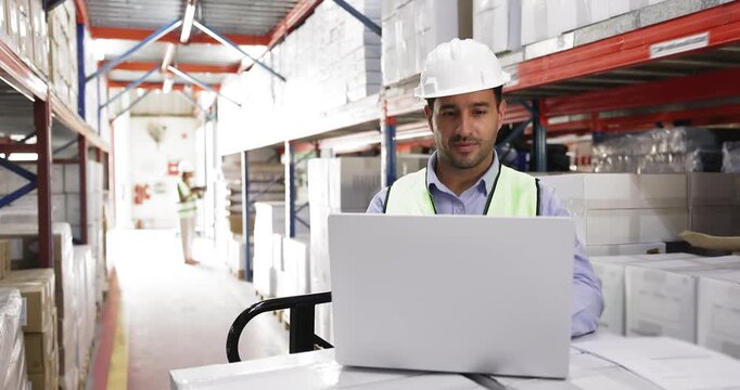 Focused Hispanic man worker wearing protective hardhat and vest using laptop, engaged in tasks related to inventory management, order processing, tracking shipments, logistics and warehouse operations