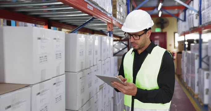 Focused Latin male logistics specialist dressed in safety vest and helmet using tablet, inspecting labelled boxes on shelves in modern storage facility, checking inventory, doing warehouse operations