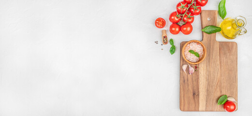 Fresh cooking ingredients with tomatoes, garlic, salt, basil, and olive oil on wooden cutting board over white background. Long banner format. top view. copy space.