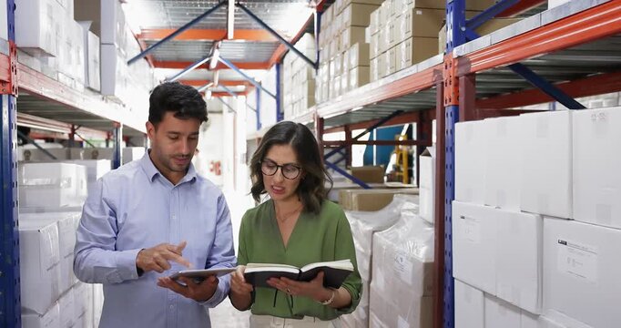 Colleagues standing in warehouse aisle surrounded by stacks of cardboard boxes, hold planner, discussing work-related matters, reviewing content on tablet. Inventory management, logistics coordination