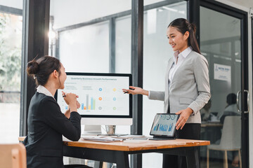 Two businesswomen in formal attire discussing quarterly business growth performance in modern office, with digital charts on computer and tablet, showing teamwork and professional collaboration