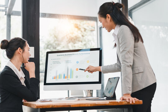 Two businesswomen in formal attire discuss quarterly business growth performance at modern office, one presenting data on computer screen with confident, positive expression - Powered by Adobe