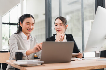 Two Asian businesswomen working together at modern office desk, discussing project on laptop with documents, coffee cup, and digital tablet, showing teamwork and focus