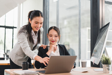 Two Asian businesswomen in formal attire working together at modern office desk, one standing and pointing at laptop screen while other sits and smiles, teamwork and collaboration