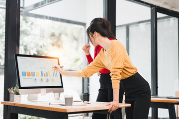 Two businesswomen analyzing data on computer screen in modern office with large windows, collaborating and discussing charts with focused expressions