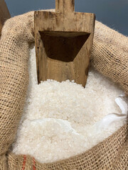 A rustic wooden scoop rests in a burlap sack filled with coarse white sea salt crystals. The contrast of rough textures creates a natural, earthy feel, evoking traditional salt harvesting methods.