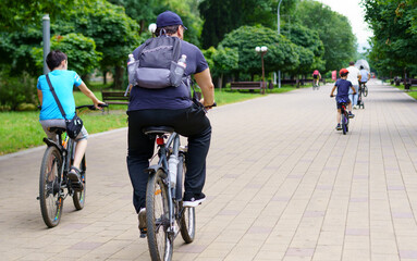 People riding bicycles through a park pathway on a sunny day with green trees and benches on either side