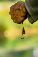 Close-up of a fisherman's hand holding a fishing lure with triple hook, set against a blurred green...