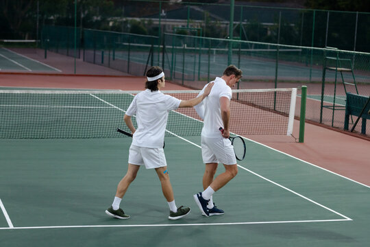 Two tennis players walking across court holding rackets near net under soft light patting shoulder