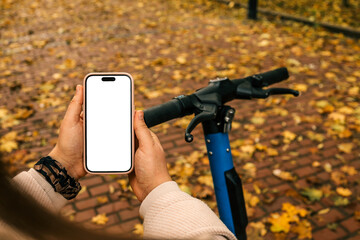 Person holding smartphone with blank screen next to electric scooter on a brick path covered with autumn leaves. Concept of navigation, mobility app, or ride-sharing in fall season