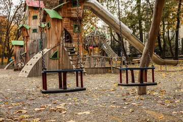 Empty swings on an autumn playground with whimsical wooden playhouses and slide surrounded by fallen leaves in the park