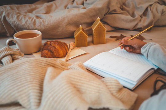 Warm autumn morning scene with a cup of tea, croissant, honey dipper, beeswax candles, cozy knit blanket, and a hand writing in a planner on a soft bed
