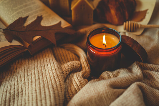 Burning amber candle with an open book, autumn leaf, croissant, and cozy knitted textures. Warm hygge atmosphere for relaxing reading time during the fall season