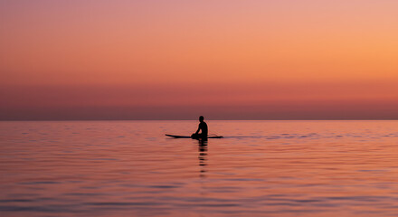 Fototapeta premium Silhouetted surfer sitting on a surfboard, waiting for the perfect waves at sunset, savoring the tranquility of the ocean and the beauty of the evening sky