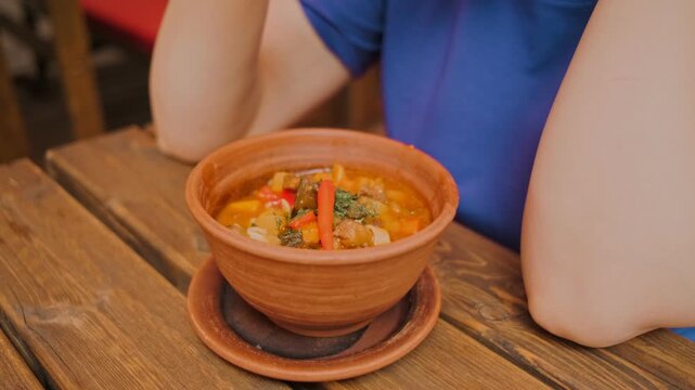 A person savors a bowl of lagman with beef featuring colorful vegetables while seated at a rustic wooden table in a charming restaurant during midday