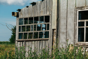 An old wooden abandoned house with broken windows stands in a field among overgrown grass on a summer day against a clear and clear blue sky with clouds. the texture of an old house