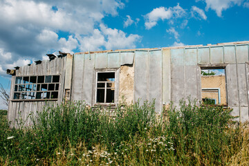 An old wooden abandoned house with broken windows stands in a field among overgrown grass on a summer day against a clear and clear blue sky with clouds. the texture of an old house