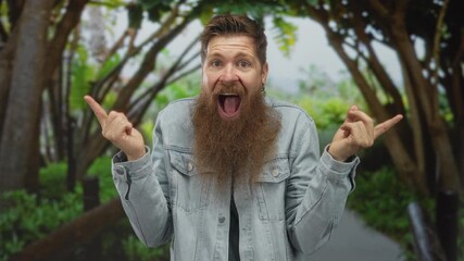 Man pointing fingers with open mouth in forest wearing denim jacket and full beard; excitement curiosity.