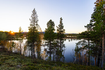 A peaceful sunset over a lake in Lapland, Sweden, with the reflection of trees in the still water.
