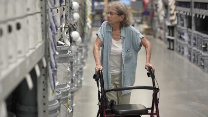 Senior woman using a walker examines various paint colors in a hardware store aisles, contemplating her options for home decoration and restoration projects.