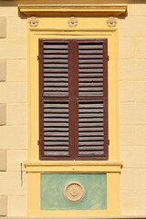 Closed, brown-painted, wood louvered shutter, yellow window-frame, cream color facade of a house on Piazza Cavour Square. Montalcino-Tuscany-Italy-135
