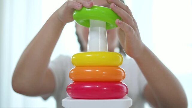 Girl playing with house on table toy. Girl carefully stacking rings on pyramid house. Girl focused on pyramid game. Educational pyramid game indoors. Brightly colored pyramid game on table. lifestyle.