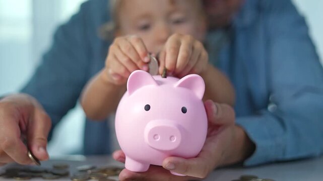 Father teaching child to save money. Child hand placing coin into piggy bank. Family saving money together. Piggy bank symbolizes financial education. Coin savings in pink piggy for child and father.