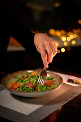 A Close-Up of a Hand Elegantly Using a Fork to Enjoy a Fresh and Vibrant Salad in a Dimly Lit Atmosphere with Warm Bokeh Lights in the Background
