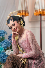 A beautiful Asian bride in traditional Javanese attire smiles gracefully, wearing a pink kebaya...