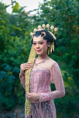 An elegant Asian woman in pink beaded kebaya poses confidently outdoors in traditional Javanese...