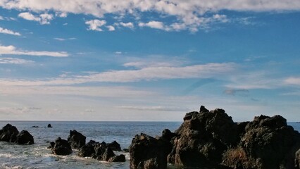 Dark coastal rocks stand amid gently swirling ocean waters beneath a bright sky with wispy clouds. A solitary rock formation dominates the foreground, evoking strength and timeless solitude.