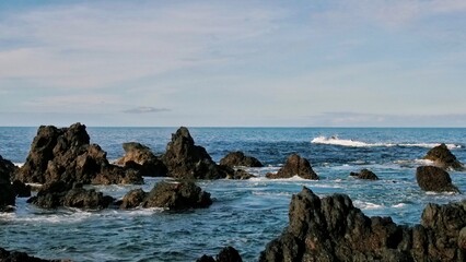 Dark coastal rocks stand amid gently swirling ocean waters beneath a bright sky with wispy clouds. A solitary rock formation dominates the foreground, evoking strength and timeless solitude.