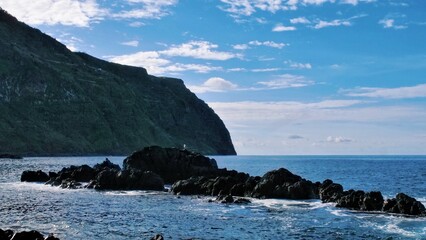Rugged coastal rocks with a perched seagull under bright skies. A dramatic cliff rises in the background, highlighting nature's contrast between wild terrain, calm ocean, and serene wildlife.