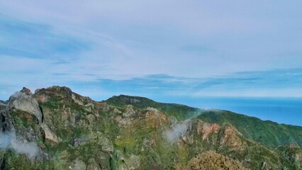Sunlit cliffs on Madeira Island rise above misty valleys, revealing reddish rock textures and vibrant green vegetation beneath a clear coastal sky. A dramatic and serene natural vista.