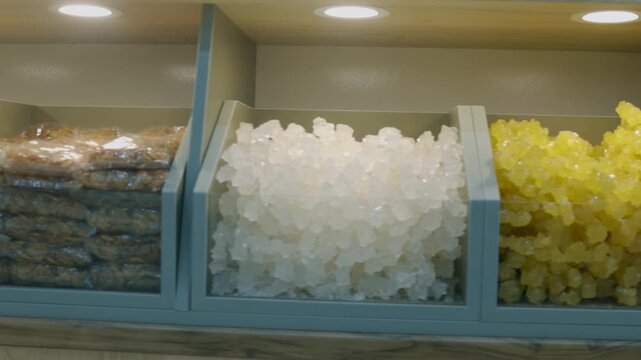 Retail shelf displaying two types of rock candy&mdash;crystal white and golden yellow&mdash;neatly arranged in plastic bins under spotlight lighting. The crystallized sugar structures are coarse and jagged