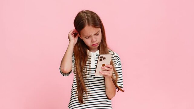 Young child girl sadly plays mobile online game on smartphone, clearly disappointed after losing a lottery jackpot and receiving bad news, showing a gloomy expression. School kid on pink background.