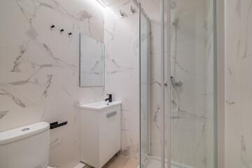 Modern bathroom with marble walls, glass shower, white sink, and toilet. Bright lighting enhances the clean, minimalist design.