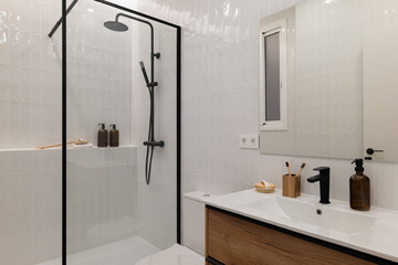 Modern bathroom with a glass shower, white tiles, wooden vanity, and minimalistic decor. Natural light enters through a window.