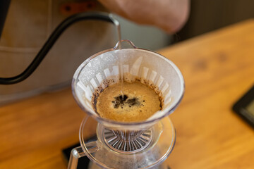 A close-up of coffee brewing in a pour-over dripper. The coffee grounds are dark and wet, with a rich brown liquid dripping into a clear carafe below.