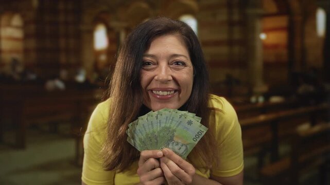 Middle-aged hispanic woman holds a fanned stack of chilean peso bills inside a vintage church nave; joy generosity hope charity.
