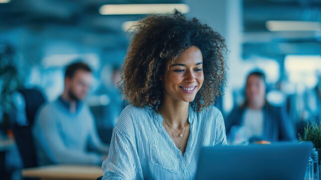 Smiling young African American businesswoman working on a laptop at her desk in a bright modern office with colleagues in the background, no logos, no brands