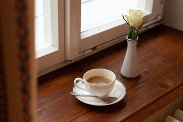 White porcelain coffee cup placed on an old wooden windowsill beside a single red rose. Cozy and nostalgic scene evoking warmth, romance, and vintage charm 